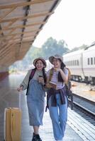 zwei jung asiatisch freunde Mädchen mit Rucksäcke beim Eisenbahn Bahnhof warten zum Zug, zwei schön Frauen Gehen entlang Plattform beim Zug Bahnhof foto