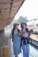 zwei jung asiatisch freunde Mädchen mit Rucksäcke beim Eisenbahn Bahnhof warten zum Zug, zwei schön Frauen Gehen entlang Plattform beim Zug Bahnhof foto