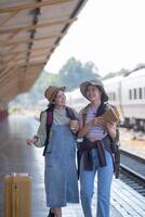 zwei jung asiatisch freunde Mädchen mit Rucksäcke beim Eisenbahn Bahnhof warten zum Zug, zwei schön Frauen Gehen entlang Plattform beim Zug Bahnhof foto