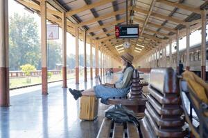 Mädchen mit ein Laptop während warten im ein Zug Bahnhof, Mädchen auf Zug Bahnhof mit Gepäck Arbeiten auf Laptop Computer, Laptop im verwenden, sitzt mit ein Koffer foto