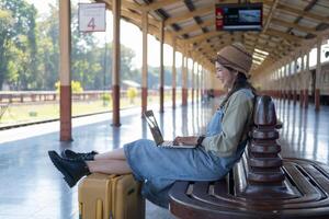 Mädchen mit ein Laptop während warten im ein Zug Bahnhof, Mädchen auf Zug Bahnhof mit Gepäck Arbeiten auf Laptop Computer, Laptop im verwenden, sitzt mit ein Koffer foto