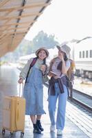 zwei jung asiatisch freunde Mädchen mit Rucksäcke beim Eisenbahn Bahnhof warten zum Zug, zwei schön Frauen Gehen entlang Plattform beim Zug Bahnhof foto