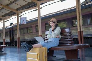 Mädchen mit ein Laptop während warten im ein Zug Bahnhof, Mädchen auf Zug Bahnhof mit Gepäck Arbeiten auf Laptop Computer, Laptop im verwenden, sitzt mit ein Koffer foto