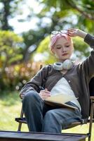 jung glücklich asiatisch Künstler Frau Zeichnung Buch und tragen Kopfhörer, das Künstler Farben im das Garten foto