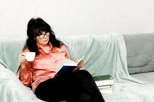 ziemlich Senior Frau auf das Sofa mit ein Tasse von Kaffee im ihr Hände lesen ein Buch foto