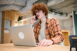 heiter männlich Freiberufler Herstellung Telefon Anruf Teilen gut Nachrichten Über Projekt Arbeiten im Cafe Innenraum, glücklich Hipster Kerl haben Smartphone Konversation während studieren im gut Stimmung Schreiben im Planer foto