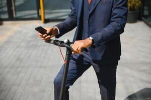 glücklich afro 30er Jahre Mann Überprüfung seine Telefon während E-Scooter Reiten im groß Stadt foto