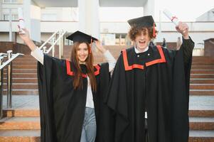 Porträt von glücklich Absolventen. zwei freunde im Abschluss Kappen und Kleider Stehen draußen Universität Gebäude mit andere Studenten im Hintergrund, halten Diplom Schriftrollen, und lächelnd foto