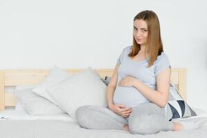 schwanger jung europäisch Frau Sitzung auf ein groß Bett und streicheln ihr groß Bauch, 7 Monat Schwangerschaft, warten zum das Geburt von ihr Baby. foto