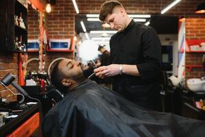 Seite Aussicht von ernst Mann mit stilvoll modern Haarschnitt suchen nach vorne im Barbier Geschäft. Hand von Barbier behalten Gerade Rasierer und Schneiden modisch Streifen auf Kopf von Klient. foto