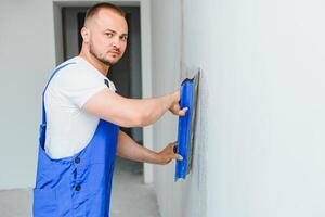 das Baumeister vorsichtig korrigiert das Unregelmäßigkeiten von das Mauer mit ein Kelle. Baumeister im Arbeit Kleider gegen ein grau Mauer. Foto Stuckateur beim arbeiten.