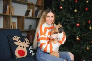 schön Frau sitzt auf ein Jahrgang Couch mit Hund. auf ein Hintergrund von ein Weihnachten Baum im ein dekoriert Zimmer. glücklich Neu Jahr foto
