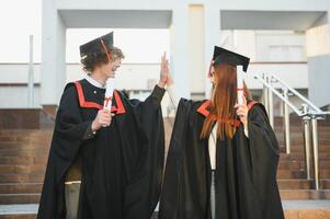 zwei froh Studenten im schwarz akademisch Kleider und Kappen Stehen im ein Bibliothek unter das Bücherregale. lehrreich Konzept. Abschluss. modern Generation. foto