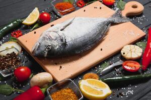 frisch roh Dorado Fisch. Meer Brachsen, Lügen auf ein hölzern Tafel auf ein Tabelle mit Kräuter und Salz- und Gewürze. Dorado und Zutaten zum Kochen auf das Tisch. Nahansicht foto