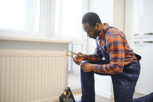Mann im Arbeitskleidung Overall mit Werkzeuge während Installation oder reparieren Heizung Kühler im Zimmer foto