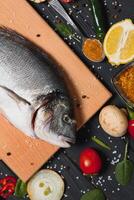 frisch roh Dorado Fisch auf Backen Papier mit Zitrone, Pfeffer, Tomaten und verschiedene Gewürze auf hölzern Hintergrund mit Kopieren Raum. foto