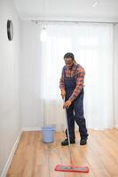 Fachmann Reiniger im Blau Uniform Waschen Fußboden und abwischen Staub von das Möbel im das Leben Zimmer von das Wohnung. Reinigung Bedienung Konzept foto