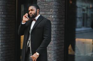 Geschäft Kommunikation. glücklich jung afrikanisch Mann im formal tragen reden auf das Handy, Mobiltelefon Telefon und lächelnd während Stehen draußen foto