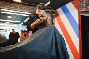 Nahansicht von Prozess von Trimmen von Haar im Barbier Geschäft. qualifiziert Barbier behalten Clipper im Hände und korrigieren gestalten von Haar zu männlich Klient Sitzung auf Stuhl. Konzept von Haarschnitt und Rasieren. foto