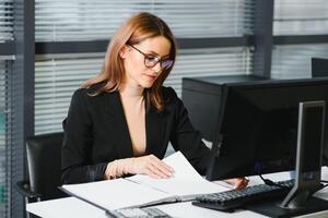 hübsch, Hübsch, Niedlich, perfekt Frau Sitzung beim ihr Schreibtisch auf Leder Stuhl im Arbeit Bahnhof, tragen Gläser, formelle Kleidung, haben Laptop und Notizbuch auf das Tabelle foto