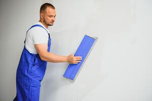 das Baumeister vorsichtig korrigiert das Unregelmäßigkeiten von das Mauer mit ein Kelle. Baumeister im Arbeit Kleider gegen ein grau Mauer. Foto Stuckateur beim arbeiten.