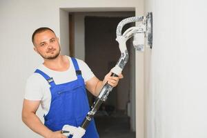 Stuckateur glättet das Mauer Oberfläche mit ein Mauer Schleifer. Meister Baumeister schleifen ein Weiß Gips Mauer. ein Mann im Overall mahlt das Oberfläche im ein Respirator. erfahren Handwerker foto