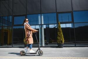 jung schön Frau im ein Jacke lächelt und Fahrten ein elektrisch Roller zu Arbeit entlang Büro Gebäude foto