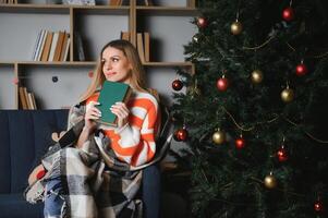 attraktiv jung Dame mit dunkel Haar lesen interessant Buch während Sitzung auf grau Couch. verwischen Hintergrund von schön Weihnachten Baum. gemütlich Atmosphäre. foto