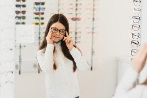 Gesundheit Pflege, Sehvermögen und Vision Konzept - - glücklich Frau wählen Brille beim Optik speichern. Porträt von schön jung Frau versuchen Neu Brille im Optiker speichern. foto