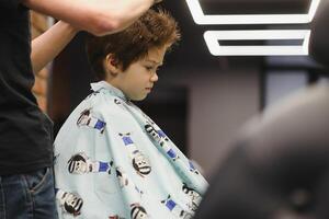 Seite Aussicht von süß wenig Junge bekommen Haarschnitt durch Friseur beim das Friseurladen. foto