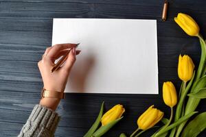 Frau ist Schreiben ein Botschaft auf das Weiß Papier. Frühling immer noch Leben. Gelb Tulpen, Weiß Papier auf das Blau Jahrgang Schreibtisch. Stift im weiblich Hand. foto