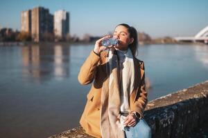 Frau genießt Trinken Wasser während Sitzung durch das Fluss auf ein sonnig Winter Tag. getönt Bild foto