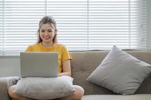 schön asiatisch Frau tragen beiläufig Kleider auf das Sofa mit ein Laptop Computer, unterhaltsam mit Sozial Medien, entspannend, lächelnd und Lachen. foto