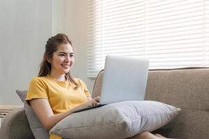 schön asiatisch Frau tragen beiläufig Kleider auf das Sofa mit ein Laptop Computer, unterhaltsam mit Sozial Medien, entspannend, lächelnd und Lachen. foto