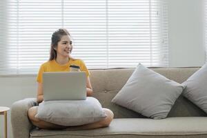 schön asiatisch Frau tragen beiläufig Kleider auf das Sofa mit ein Laptop Computer, unterhaltsam mit Sozial Medien, entspannend, lächelnd und Lachen. foto