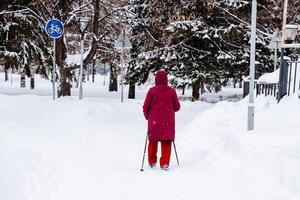 ein Skifahrer im ein hell rot passen Gehen im Stadt Park. Rennen Gehen mit Stangen. Oma Theaterstücke draussen Sport. gehen im das frisch Luft foto