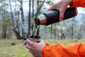 das Konzept von heiß Getränke im kalt Wetter im Natur, ein des Mannes Hände dauerte ein Thermosflasche, ein Kerl gießt Tee in ein Glas, Camping im das Wald zu trinken ein heiß trinken, Erwärmen überlegte Wein foto