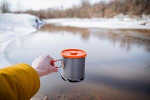 Hand hält ein Topf gegen das Hintergrund von Wasser, Geschirr im ein Wanderung schwenken mit ein Deckel, ein Licht Topf zum Essen, ein Kühler beim das Unterseite von das Pfanne, Ausrüstung zum ein Tourist. foto