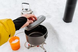 ein des Mannes Hand hält ein Deckel von ein Topf, ein grau Topf steht auf das Feuer, ein Wanderer Wanderung Köche Essen auf ein Gas Camping Brenner, Kletterer Gang, Trekking Winter Mittagessen foto