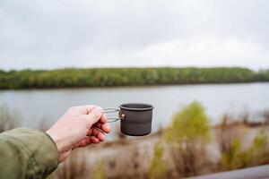 Kaffee Becher zum Camping im Natur, Mann Hand halten ein Becher, Tee Glas, Metall Tasse zum heiß Getränke, Aluminium Utensilien foto