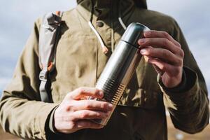 Vakuum Silber Thermosflasche ist gehaltenen im das Hände von ein Mann, das Konzept von ein Camping Küche, ein Metall Flasche zum heiß Getränke, Ausrüstung zum ein Wanderung. foto