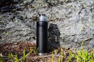 schwarz Thermosflasche steht auf das Hintergrund von ein Stein, Vakuum Flasche zum Kaffee, ein Schiff zum ein heiß trinken, Nein Deckel, Grün Gras, ein Stück von Felsen, Metall Stahl foto