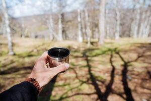 ein Becher im das Hand von ein Person gegen das Hintergrund von das Wald, ein Tasse von heiß Kaffee früh im das Morgen im Natur im das Wald, ein Armband von Rudraksha auf das Handgelenk, ein Wanderung im das Berge foto