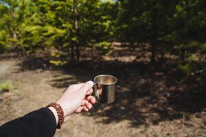 ein des Mannes Hand hält ein Becher von Tee gegen das Hintergrund von das Wald, ein Metall Becher zum Kaffee zu trinken auf ein Wanderung, Geschirr im das Wald, Natur Kiefer Sonne, Rudraksha Armband. foto