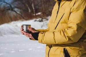 ein Glas im Hand. ein Becher von heiß Tee, ein Mann halten ein Tasse von heiß trinken, ein Gelb Jacke, ein warm Nieder Jacke, Winter. foto