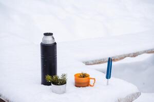 ein schwarz Thermosflasche und Tassen mit el's Zweig im das Schnee. ein Wandern Messer werden Sein stecken im das Schnee. Winter immer noch Leben im das Schnee foto