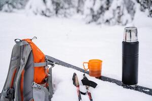 ein schwarz Thermosflasche mit ein Becher von Tee Stand auf Ski im das Schnee. ein hell Orange Wandern Rucksack ist Lügen im das Schnee. Skifahren im Winter. foto