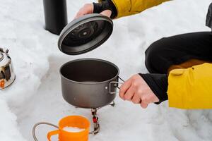Kochen im Natur beim das Campingplatz, Winter Wandern schwenken steht auf das Tourist Brenner, Hände nehmen ein Topf von Essen, Kochen im das Schnee extrem Winter foto