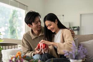 romantisch jung asiatisch Paar Umarmen geben Geschenk im Leben Zimmer beim heim. fallen im Liebe. Valentinstag Konzept foto