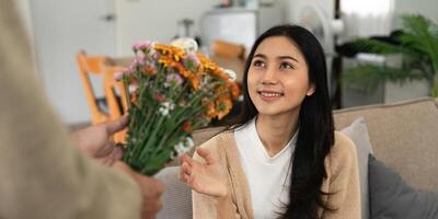 romantisch jung asiatisch Paar Umarmen mit halten Blumen und lächelnd im Leben Zimmer beim heim. fallen im Liebe. Valentinstag Konzept foto